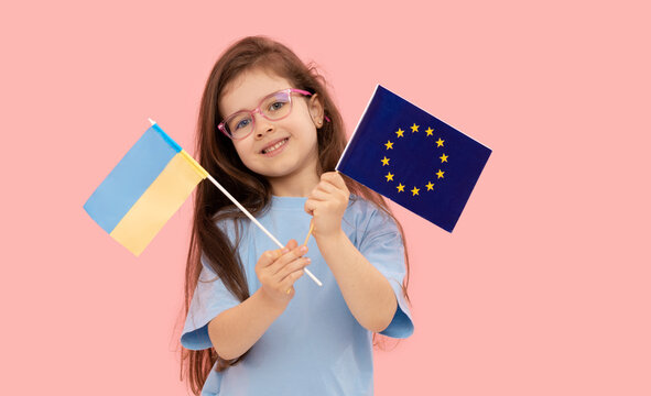 Cute child girl holding in her hand the flag of the European Union and Ukraine on a pink background. The concept of supporting the union of states in war