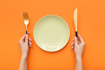 Female hands with cutlery and empty plate on orange background
