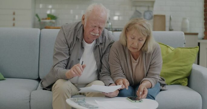 Stressed retired people counting rent and household payments paying bills using smartphone application at home. Retirement and finance concept.