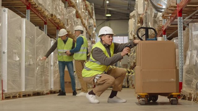 Biracial female warehouse worker in hard hat and acid green vest scanning barcodes on cardboard boxes loaded onto hand pallet truck