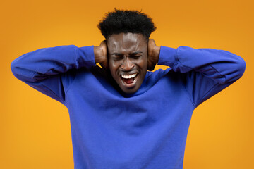 Young african man screaming, covering ears on yellow