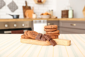 Wooden board with cookies, chocolate and glass of milk on table in kitchen
