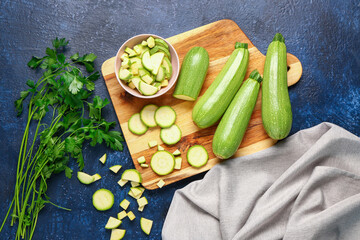 Board with fresh green zucchini and bowl of slices on blue background