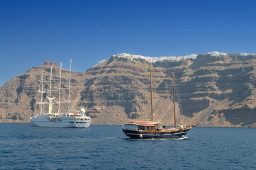 Retro schooner and modern sailing ship floating off the rocky coast of Santorini island.