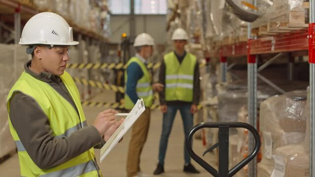 Medium shot of young female warehouse worker signing invoice document while doing inventory management during shift