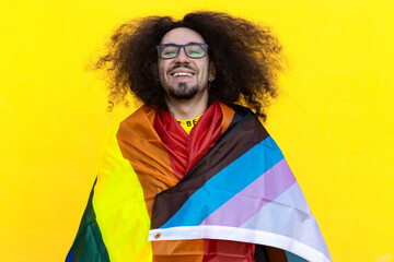 portrait of a happy man with glasses beard and long hair wrapped in a transgender LGBT flag isolated on yellow background