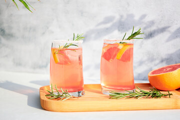 Chilled summer cocktails of grapefruit or red orange, rosemary and ice on a wooden serving tray on a gray background on a sunny sunny day. Vacation concept.