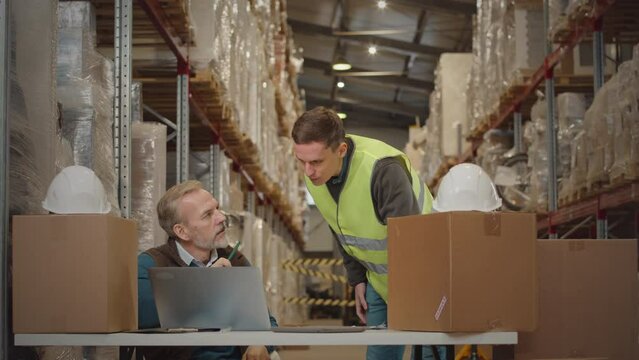 Medium shot of mature warehouse manager speaking to young employee while doing inventory of goods packed in cardboard boxes