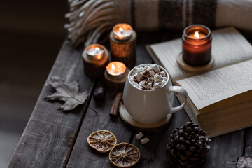 Autumn cozy home composition with hot chocolate with marshmallow and candles. Aromatherapy on a grey fall morning, atmosphere of cosiness and relax. Wooden background, books, close up.