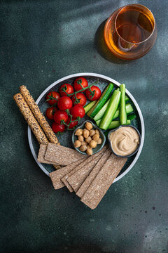 Overhead view of a plate of hummus, chickpeas, crispbread, breadsticks, tomatoes and cucumber batons with a glass of wine