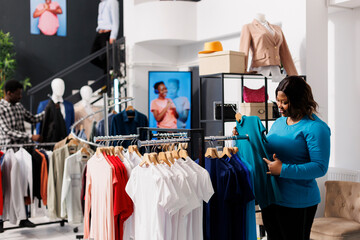Shopaholic african american woman checking elegant blue shirt, checking formal wear fabric in clothing store. Stylish customer shopping for fashionable merchandise and accessories in modern boutique.