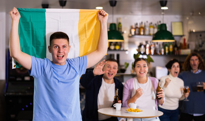 Company of emotional young adult fans supporting favorite team with flag of Ireland while resting...