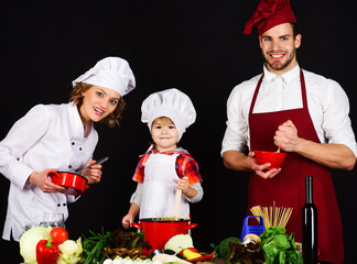 Happy family in kitchen preparing healthy food. Adorable son in chef hat with parents cooking together homemade food. Preparation to breakfast, dinner or supper. Parents teaching little boy to cook.