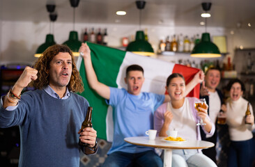 Company of screaming young adult sports fans waving flag of Italy and supporting national team with...