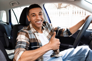 Happy black man sitting in new car and showing thumbs up sign gesture, buying modern vehicle in...