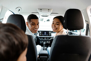 Back view of african american young family of three sitting in new car looking at child and smiling, enjoying summer road trip together