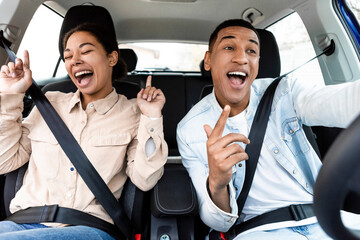 Joyful black lady and man singing, dancing and enjoying travel in car together, being in good mood,...