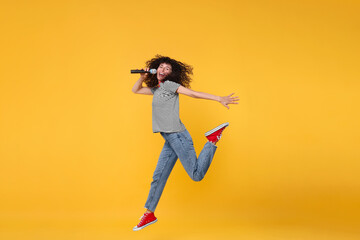 Beautiful young woman with microphone singing and jumping on yellow background