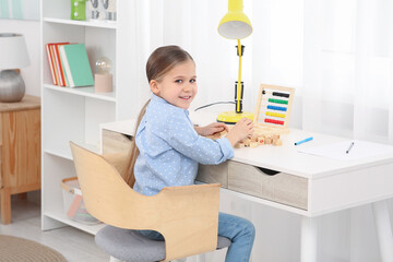 Cute little girl playing with wooden cubes at desk in room. Home workplace