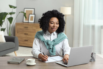 Happy young woman writing notes while using laptop at wooden desk indoors