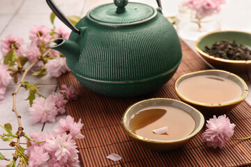 Obraz na płótnie Canvas Traditional ceremony. Cup of brewed tea, teapot and sakura flowers on bamboo mat, closeup