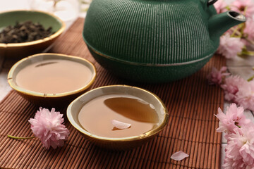 Fototapeta na wymiar Traditional ceremony. Cup of brewed tea, teapot and sakura flowers on bamboo mat, closeup