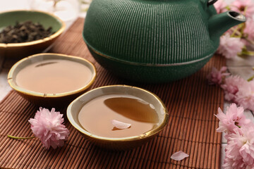 Traditional ceremony. Cup of brewed tea, teapot and sakura flowers on bamboo mat, closeup