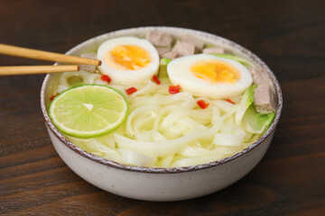 Bowl of delicious rice noodle soup with meat and egg on wooden table, closeup
