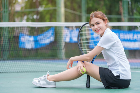 Female tennis player in sports skirt sitting by the net on court, holding a rocket and smiling to camera.