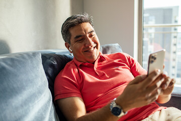 MIddle aged latin man using a smart phone on a couch at home © Baba Images