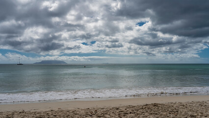 Serene seascape. The silhouette of a yacht is visible in the boundless turquoise ocean. The island on the horizon is hiding in the clouds. Waves are foaming on the sandy beach. Seychelles. Mahe. 