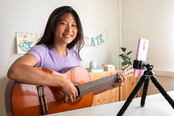 Young Asian woman recording herself playing guitar with phone. Social media live stream. Asian girl...