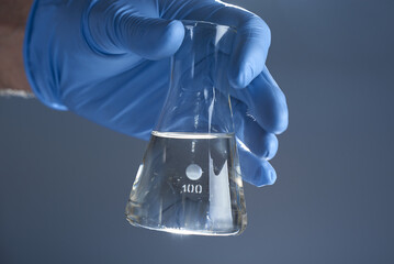 A hand in a medical glove holds a laboratory flask of liquid on a gray background. The concept of a...