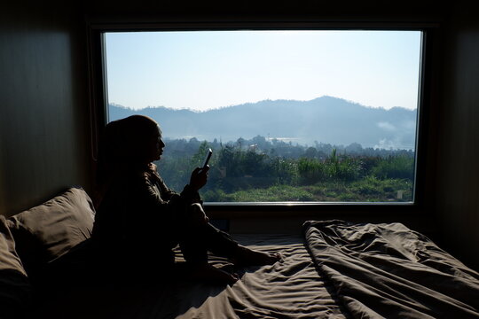 Sad woman is sitting on bed and looking at beautiful landscape behind glass window.