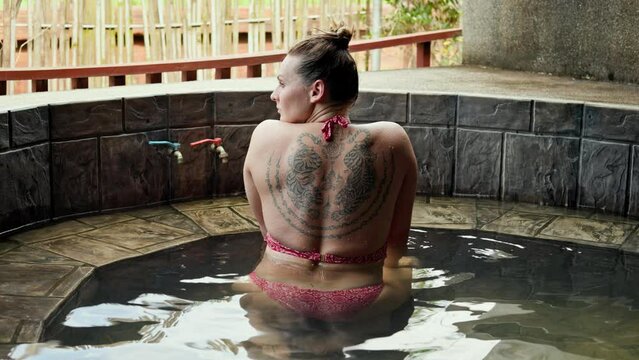 Woman with athletic body and sak yant tiger tattoo on her back sits in a hot pool