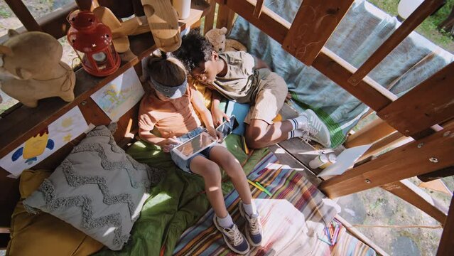 Overhead shot of African American and Caucasian elementary age boys lying in treehouse playing games in tablet
