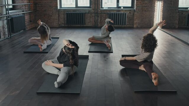 Wide shot of four diverse women sitting on mats in loft studio doing stretching asana during yoga workout