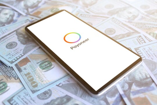 Payoneer logo displayed on smartphone on top of pile of money. Business profit concept