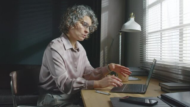 Medium portrait of female psychotherapist sitting at desk in front of laptop talking to her patient during online consultation