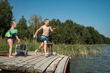 Fototapeta na wymiar cute caucasian boy and girl jumping from wooden pier diving into lake in countryside
