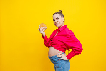 Funny pregnant young woman eating burger. Pregnant woman holding tasty sandwich while standing isolated on yellow studio wall. Junk food concept. Fast food during pregnancy.