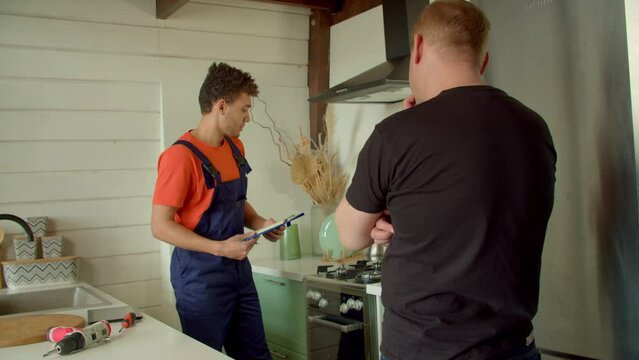 Skillful attractive black male technician in protective workwear telling customer about repair service work, showing clients fixed kitchen appliance and writing done into clipboard indoors.