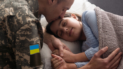 Soldier in Ukrainian military uniform kissing his daughter while she sleeping on sofa at home....