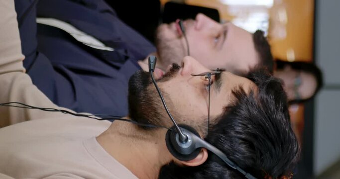 Vertical shoot. Adult handsome office worker wearing headhpones with microphone satisfait his job. Berded man focused on work is sitting at workplace. Operator of hot line.