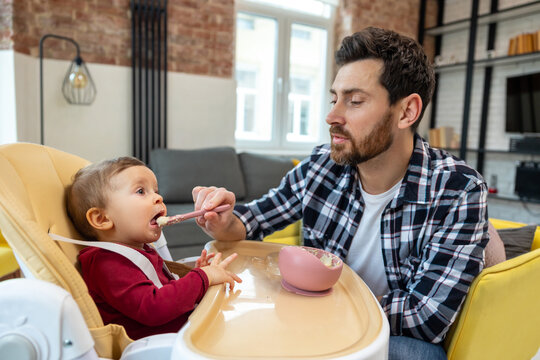 Bearded father feeding little baby daughter sitting in highchair with puree or porridge.