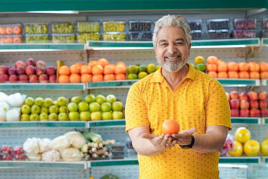 Senior indian man showing orange fruit at fruit shop. Healthy lifestyle concept.