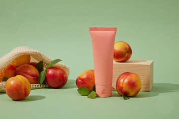 Front view of a pink plastic tube unbranded decorated with fresh ripe peaches (prunus persica) and wood block on a pastel background. Mockup for cosmetic from peach ingredient and space for design