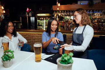 Happy young women in casual clothing paying for drinks with card machine next to waitress