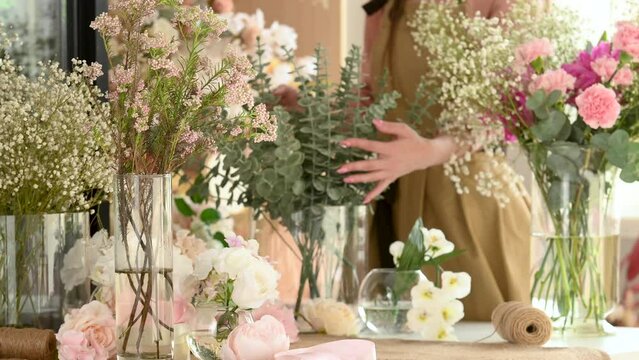 Positive young female florist in green apron standing at table and cutting flowers in floral shop. Woman florist makes a bouquet at home in the decoration studio. Flowers and vases close-up.