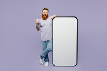 Full body fun young redhead man wear violet t-shirt casual clothes big huge blank screen mobile cell phone smartphone with copy space mockup area do winner gesture isolated on plain purple background.